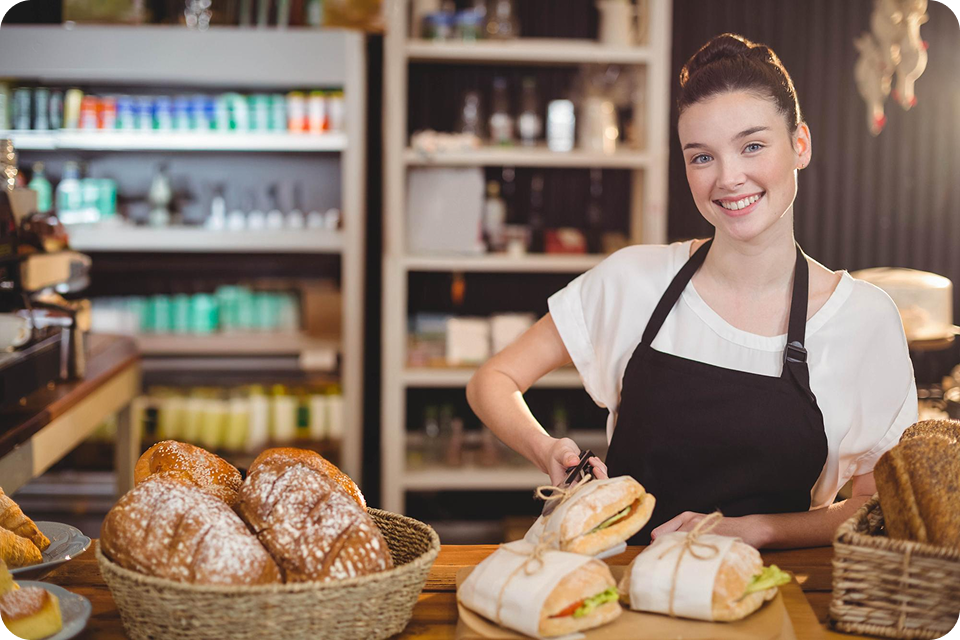 A woman in an apron stands behind a counter in a bakery/café, surrounded by fresh food. It represents F&B (food and beverage) SMEs. ccMonet’s AI - powered tools (bookkeeping, payroll) and industry - tailored services, plus expert support (tax compliance, business registration), help such businesses manage finances efficiently.